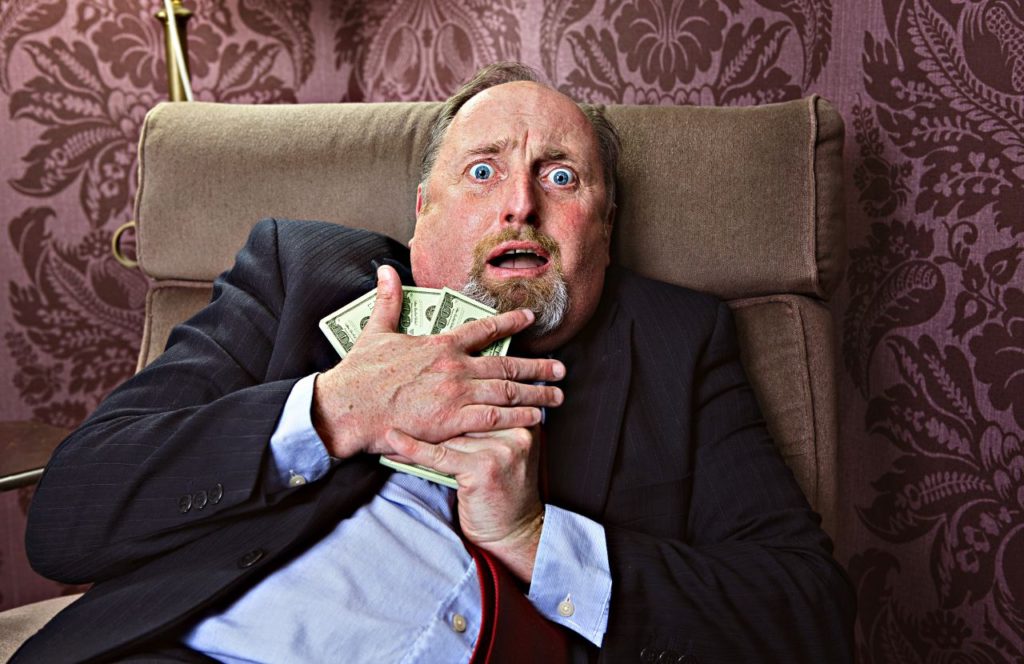man in a suit sitting in a chair with big eye holding hundred dollar bills which can be an example of tax evasion vs tax fraud