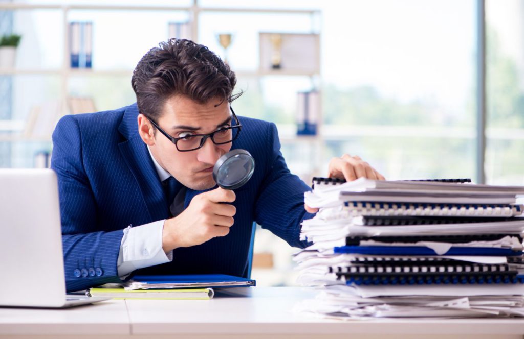 man with glasses looking through a magnifying glass inspecting a stack of documents