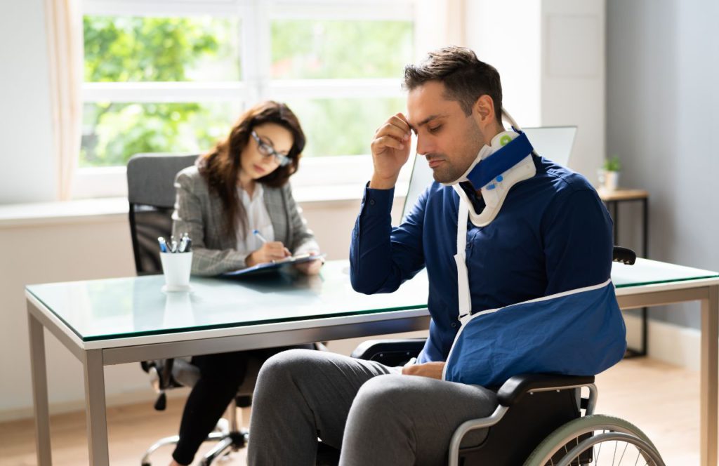 man in a wheelchair with neck brace with a woman at a desk filling out his car accident settlement