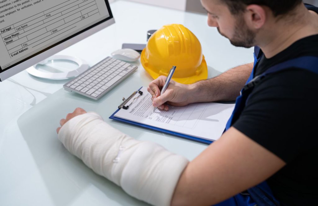 white man with cast on left arm filling out his car accident settlement form in front of a computer with a yellow helmet on the right side.